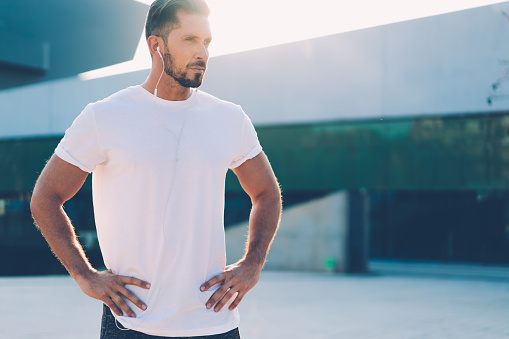 Caucasian male in earphones and blank t-shirt with copy space for brand name or label listening sportive music podcast, muscular athlete 30 years old standing at urban street planning workout