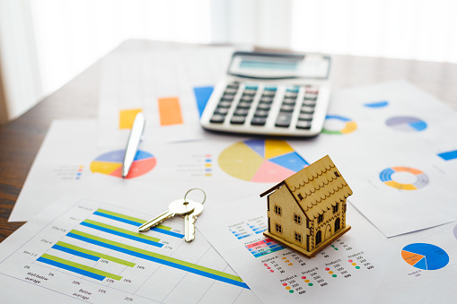 Color image depicting a real estate market and property concept. A wooden model house, a set of house keys, a calculator and pen sit on top of an array of documents with financial bar graphs and pie charts.