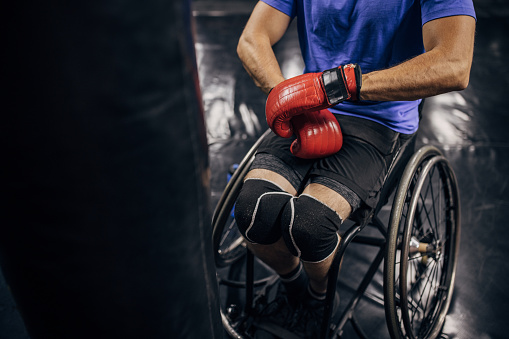 One man, unrecognizable disabled male in wheelchair putting on boxing gloves in gym.