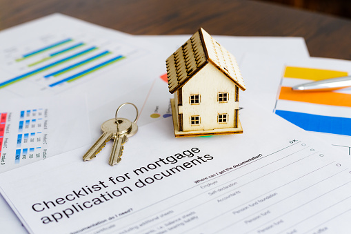 Color image depicting a real estate market and property concept. A wooden model house, a set of house keys, a calculator and pen sit on top of an array of mortgage documents with financial bar graphs and pie charts.