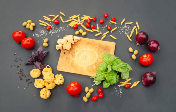 Cutting board, fresh tomatoes and onions, raw penne and fettuccine, cheese, basil, green olives and spices on dark background. Top view, copy space.