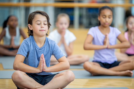 A group of school aged children sit out on yoga mats in the gymnasium as they stretch and meditate together.  They are each dressed comfortably in athletic wear and are sitting with their eyes closed as they focus on their breathing.