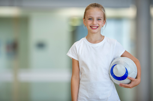 A young school aged girl stands in a gymnasium as she poses for a portrait.  She is dressed comfortably in athletic wear and is holding a volleyball on her hip as she smiles.