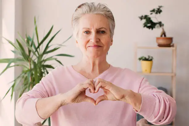 Cute happy middle aged mature woman making heart shape hand gesture looking at camera, funny smiling adult single senior woman blogger laughing face showing love sign symbol, dating concept, close up portrait