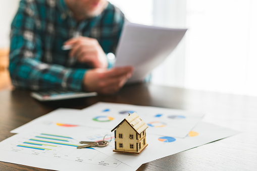 A man looking at documents pertaining to a house sale agreement. Focus on a model house on top of documents in the foreground, while the man - sitting at a table and studying the agreement papers - is defocused beyond.