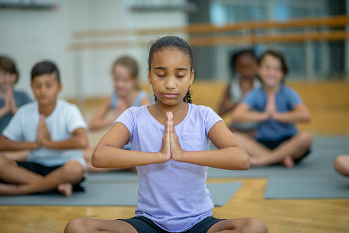 A group of school aged children sit out on yoga mats in the gymnasium as they stretch and meditate together.  They are each dressed comfortably in athletic wear and are sitting with their eyes closed as they focus on their breathing.