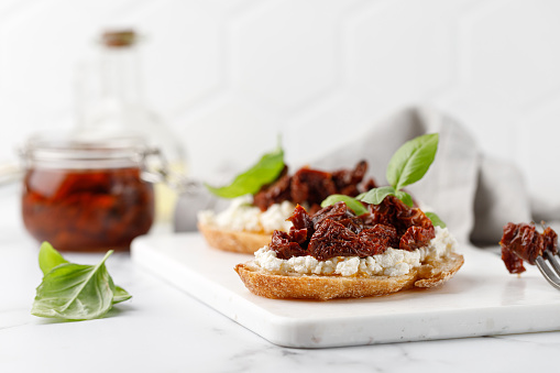 Sandwiches with dried tomatoes in oil, cottage cheese, ricotta cheese, and basil. Bruschetta, snack, appetizers. Toasts on white board and marble background.
