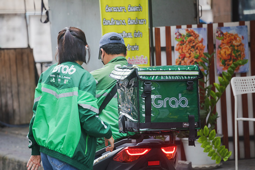 Thai couple with  motorcycles and  GRAB delivery box is standing in front of a street food reataurant in residential district of Bangkok Chatuchak. Man is using mobile phone. Both are wearing protectuve face mask. Scene is near CRU university