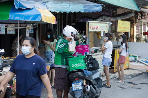 Thai GRAB delivery is taking a selfie with food bag in street. Man is standing in front of street food market stalls of local market. Scene is near CRU university in residential district of Bangkok Chatuchak. Some people are walking in scene. People are wearing protective face masks