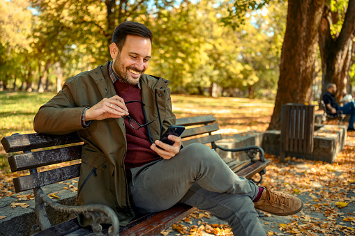 Smiling man sitting at park bench and using smartphone