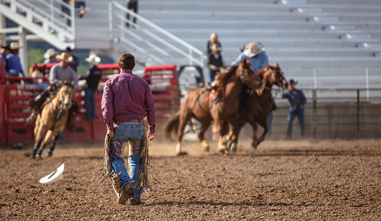 A male rodeo rider wearing a purple shirt and blue jeans walks towards a small group of horseback riders riding in front of him.