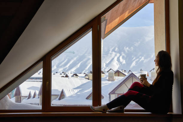 Woman being cold sits on windowsill, warming up with tea mug, looking at snow covered mountains. Female drinks coffee in cozy chalet wearing thermal underwear layer. Rising heating bills on winter. stock photo