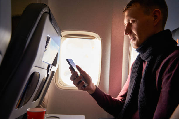 istock Businessman sitting next to airplane window and using smartphone. Man fly on plane and reading with phone in online app. Person using internet on air flight. Guy booking hotel online. 1434486899