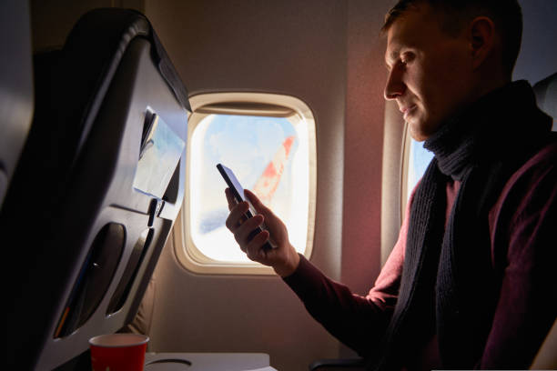 istock Businessman sitting next to airplane window and using smartphone. Man fly on plane and reading with phone in online app. Person using internet on air flight. Guy booking hotel online. 1434487294