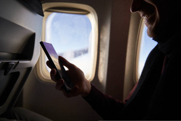 istock Businessman sitting next to airplane window and using smartphone. Depersonalized man fly on plane and reading with phone in online app. Person using internet on air flight. Guy booking hotel online. 1434487771