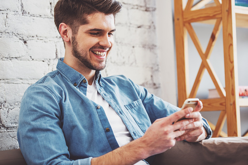 Attractive young man is using a smartphone and smiling while sitting on sofa at home