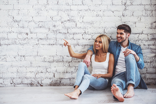 Beautiful young couple is hugging and smiling while sitting on floor against white brick wall. Girl is pointing away