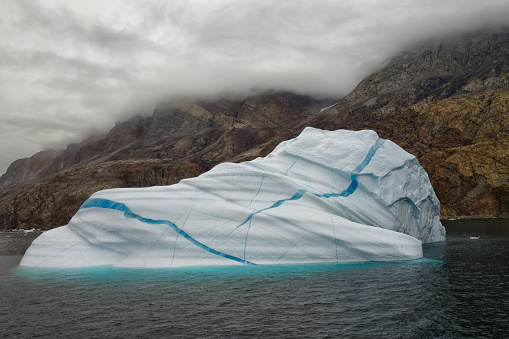 A large iceberg is seen in a Greenland fjord.  The iceberg is large and blue in color.  Mountains can be seen in the background.  The iceberg has many lines of deep blue ice in them.  The pattern caused by the glacial lines is very dramatic