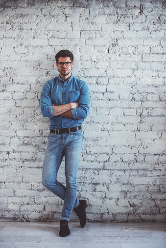 Full lenght portrait of handsome young man in jeans clothes and eyeglasses looking at camera and smiling, standing against white brick wall
