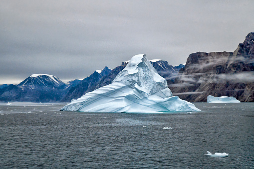 A group of icebergs is seen at dusk.  A foggy mist partially covers the icebergs.  2 large mountains can be seen in the background.  The mountains are rugged and of different colors.  This photo was taken in a Fjord in Greenland