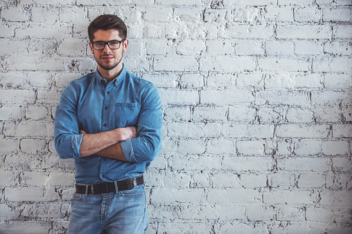 Portrait of handsome young man in jeans clothes and eyeglasses looking at camera and smiling, standing against white brick wall