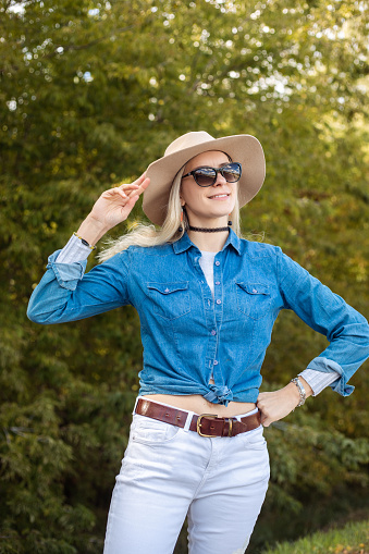 Portrait of young wonderful blond woman wearing blue shirt, white jeans, sunglasses, touching beige floppy hat, standing near green trees forest park. Travelling, hitchhiking, adventure, summer.
