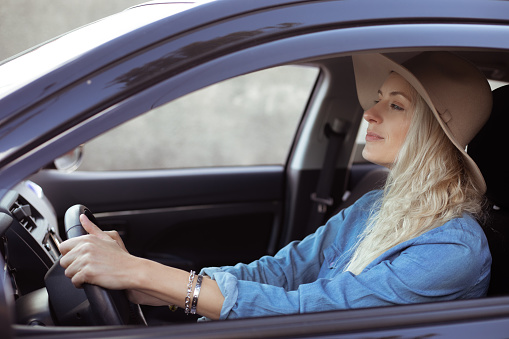 Portrait of young cheerful beauteous woman with long fair hair wearing blue denim shirt, beige floppy hat, sitting in black car, holding steering wheel, driving. Travelling, adventure, transport.