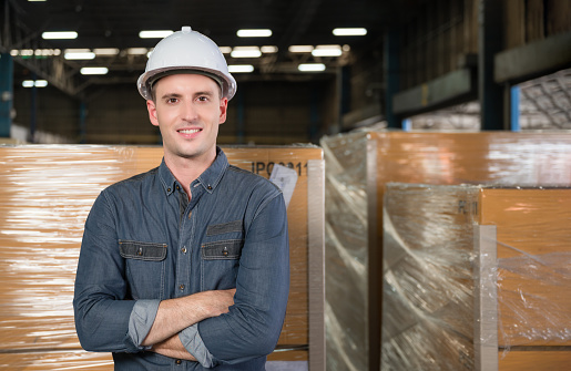 Portrait of happy professional handsome engineer man wearing safety white helmet standing arms crossed in logistics warehouse. Confident male business manager smiling and looking at camera in factory.