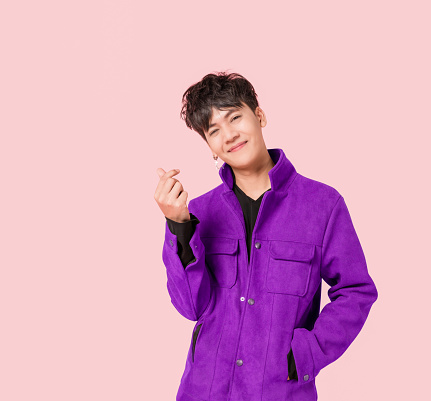 Retrato de un hombre guapo asiático feliz con ropa de chaqueta púrpura de moda de pie sonriendo con los dedos para hacer un mini signo de corazón aislado sobre fondo rosa. photo