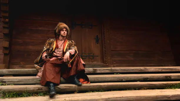 Pensive young ukrainian cossack dressed traditional clothing holding saber in his hand sitting on the steps and looking into the distance on background of old wooden door