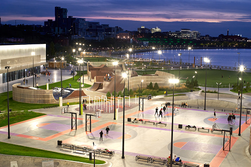 Evening above view of the illuminated playground at city embankment. Rare unrecognizable people in the illuminated skatepark. Blue sky and river, green grass. Kazan city, Russia
