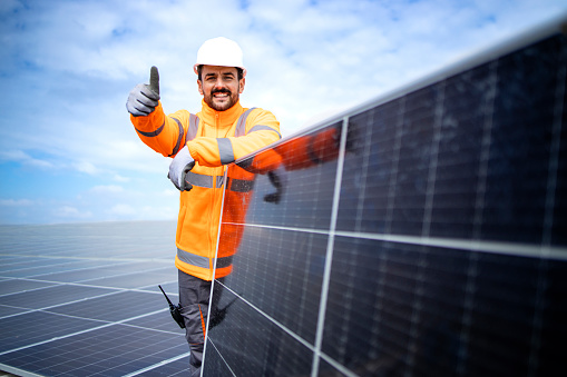 Portrait of professional worker standing by solar panel and holding thumbs up. Installation of solar power plant. Sustainable energy source.