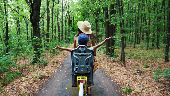 a woman in a hat and dress, with a basket of flowers, together with small child, ride bicycle, in the forest, in the summer. the child is sitting in a special chair. High quality photo