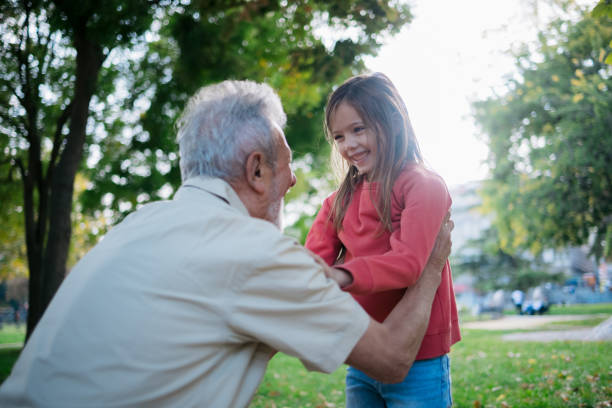 Happy grandfather enjoying himself outdoors with his granddaughter in the public park, holding her in his arms and bonding with her