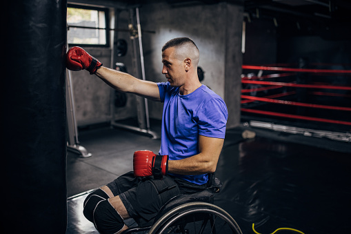 One man, disabled male boxer in wheelchair training with punching bag in gym.