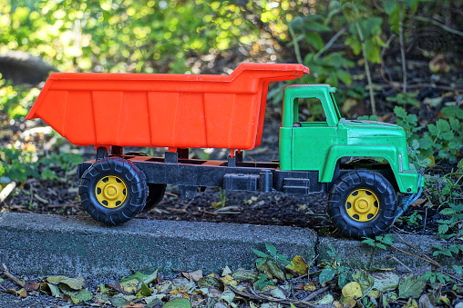 one large plastic toy car truck with a green cab and a red body stands on a gray concrete curb in the street among the vegetation