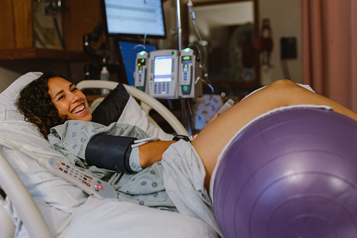 A Eurasian pregnant woman laughs while lying in a hospital bed in a labor and delivery room. The woman is using a peanut ball to help open her pelvis so labor progresses faster and to aid with pain management during contractions.