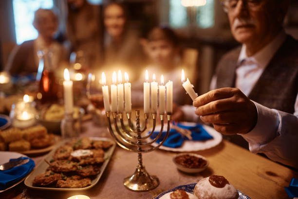 Close up of senior man lighting menorah during family dinner on Hanukkah. Close up of extended Jewish family celebrating Hanukkah at dining table. Focus is on mature man lighting candles in menorah. orthodox judaism photos stock pictures, royalty-free photos & images