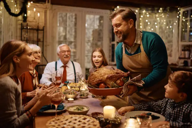 Happy man bringing roast turkey at the table during Thanksgiving dinner with his multigeneration family.