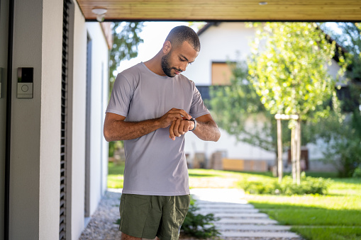 Young african american sportive man in wireless earphones looking at smart watch on wrist, medium shot. Person checking fitness tracker before training outdoors, side view