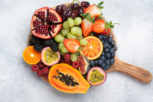 Delicious fruit on round wood chopping board, mango pomegranate raspberries papaya oranges passion fruits berries on off white concrete background, selective focus