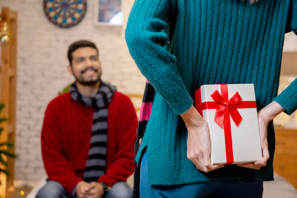 Rear view young caucasian woman hide presents for surprise boyfriend in celebration Christmas at home, woman holding gift hide behind for giving man with excited, xmas and new year or holiday concept. stock photo