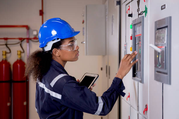Electrical young asian woman engineer examining maintenance cabinet system electric and using tablet in control room at industrial factory, technician or electrician inspection power distribution. stock photo