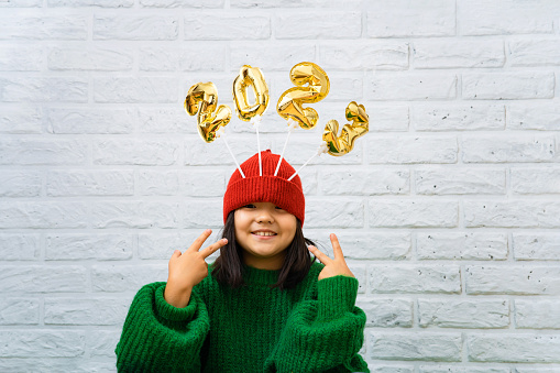 Happy Asian baby girl in a green sweater close-up with golden balloons with numbers 2023 in a hat. Copy space, white brick wall background, girl looking at the camera, smiling