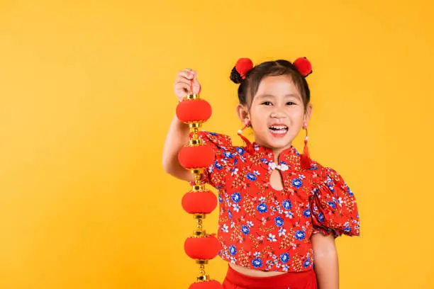 Chinese New Year. Happy Asian Chinese little girl smile wearing red cheongsam qipao holding silk lanterns on hand, Portrait children in traditional dress, studio short isolated on yellow background