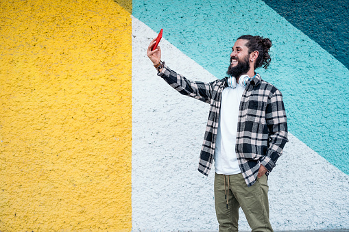 Happy hipster man standing against colorful wall in the city and taking a selfie with his phone.
