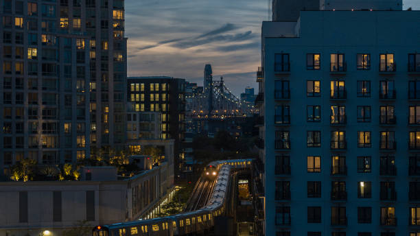 zug fährt auf der erhöhten u-bahn-linie zwischen gebäuden in long island city, queens bei nacht mit queensboro bridge dahinter. - housing development development residential district aerial view stock-fotos und bilder