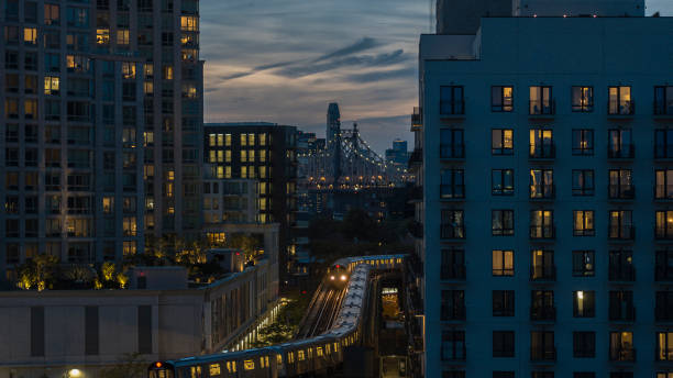 Train riding on the elevated subway line between buildings in Long Island City, Queens at night with Queensboro Bridge seen behind. stock photo