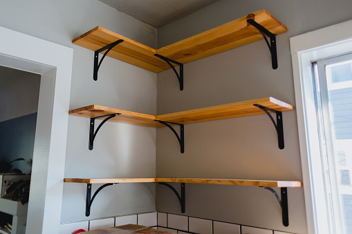 Three open faced shelfs in a home kitchen. These shelfs warp around a corner and are wood finished texture with metal brackets. They are mounted between a door frame and window frame.