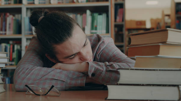istock Handsome young man sitting with his head on table is sleeping in school library surrounded by huge piles of books. His glasses are lieing in front of him 1434616813